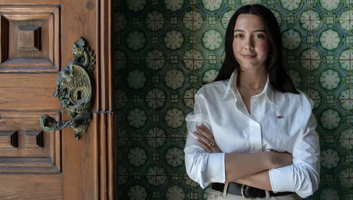 Foto de Leonor Caldeira. A Leonor aparece a meio corpo, a olhar directamente para a camera, com um sorriso confiante e uma branco, contra um fundo de azulejo e ao lado de uma porta de madeira antiga.
